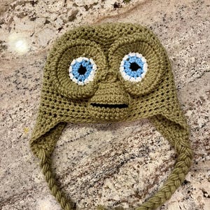 May include: A handmade olive green crochet hat shaped like an alien's head, featuring large eyes with blue centers and white outlines. The hat has a detailed face and braided ties with tassels.
