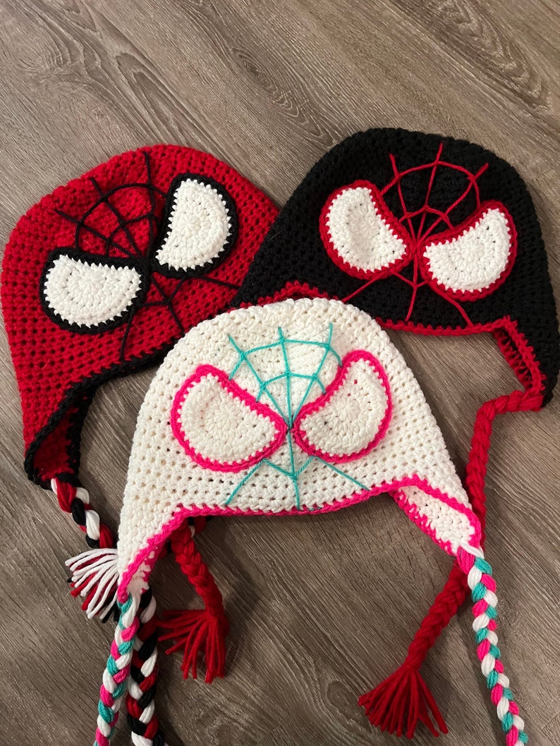 May include: Three crocheted hats with Spiderman designs. The hats are red, black, and white with white eyes and a red spider web design. The hats have long braided ties.