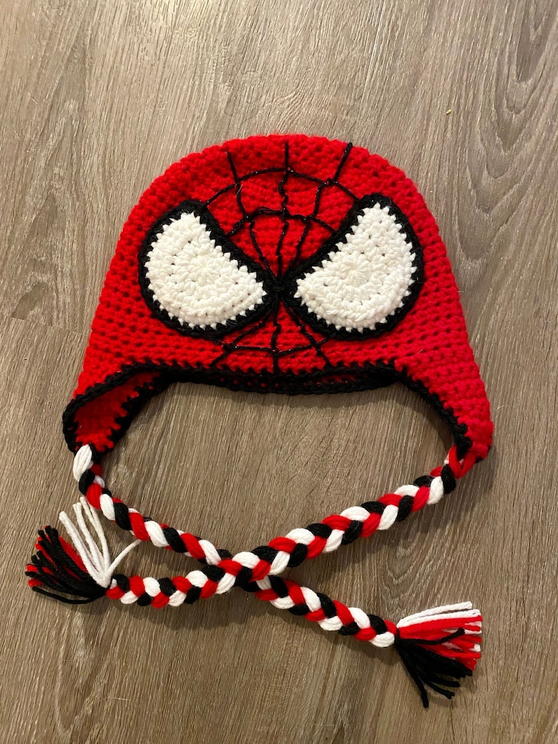 May include: Red crocheted hat with a black spider web design and white eyes. The hat has a long, braided cord with red, white, and black yarn.