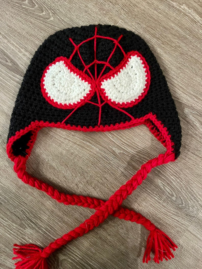 May include: Black crochet hat with a red and white spider-man design. The hat has a long red braided cord with tassels.