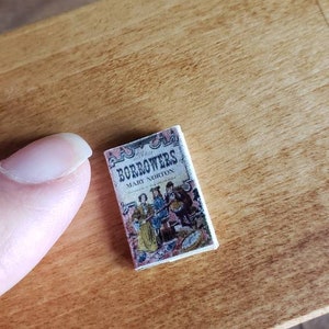 May include: A miniature hardcover book titled "The Borrowers" by Mary Norton. The cover features an illustration of four small people in a kitchen setting.