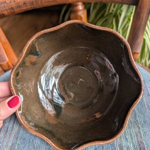 May include: A handmade, dark brown ceramic bowl with a wavy rim. The bowl has a glossy finish and a slightly textured surface. The rim is a lighter brown color. The bowl is sitting on a blue and green patterned fabric.