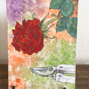 May include: A vibrant painting of a red rose with green leaves and a pair of silver pruning shears. The background features a blend of orange, green, and purple hues, creating a textured effect. The artwork is displayed on a black metal stand.