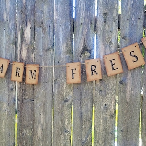 May include: A rustic brown paper banner with the words "FARM FRESH" printed in black. The banner is strung on a twine string and hung on a wooden fence.