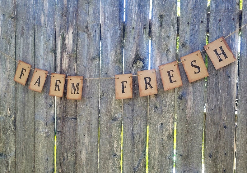 May include: A rustic brown paper banner with the words "FARM FRESH" printed in black. The banner is strung on a twine string and hung on a wooden fence.