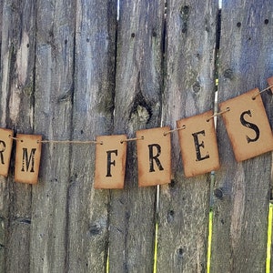May include: A rustic brown paper banner with the words "FARM FRESH" strung on twine against a wooden fence background.