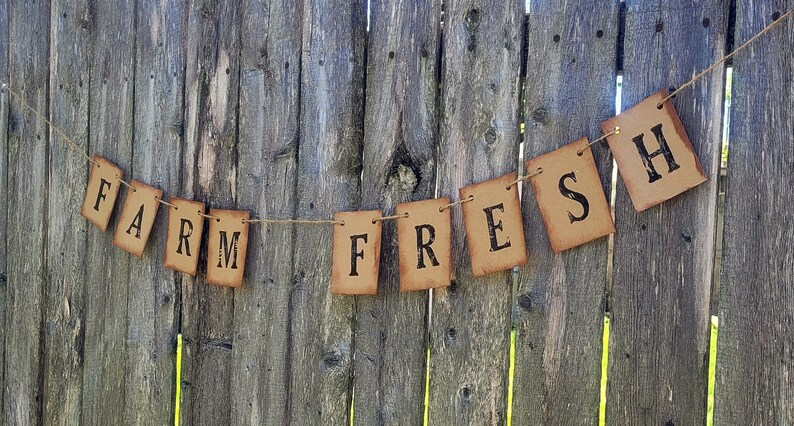 May include: A rustic brown paper banner with the words "FARM FRESH" strung on twine against a wooden fence background.