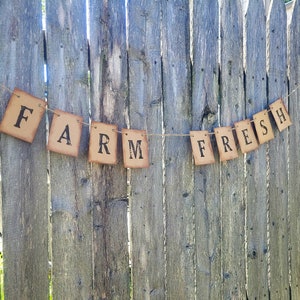 May include: A rustic brown banner with the words "Farm Fresh" strung on twine against a wooden fence background.