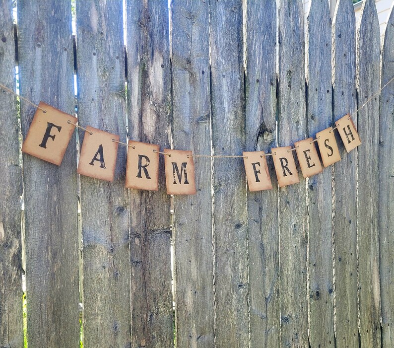 May include: A rustic brown banner with the words "Farm Fresh" strung on twine against a wooden fence background.
