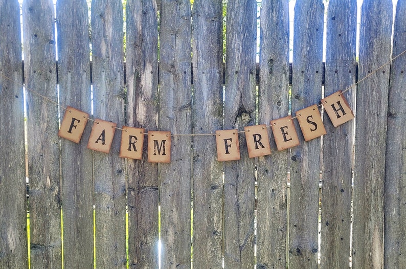 May include: A rustic brown paper banner with the words "FARM FRESH" strung on twine against a weathered wooden fence.