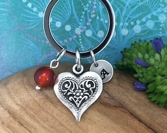 May include: Silver-tone keychain with a decorative heart pendant, a red bead, and a small disc with the letter "A". The heart pendant has an ornate design. The keychain is a personalized accessory.