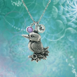 May include: Silver necklace featuring a detailed bird charm, a purple gemstone, and a round charm with the letter "R" on it.