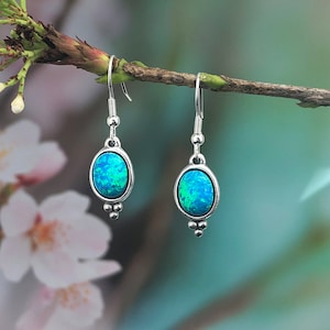 May include: A pair of silver dangle earrings featuring oval-shaped blue opal stones. The earrings have a simple design with three small silver beads at the bottom of each earring.