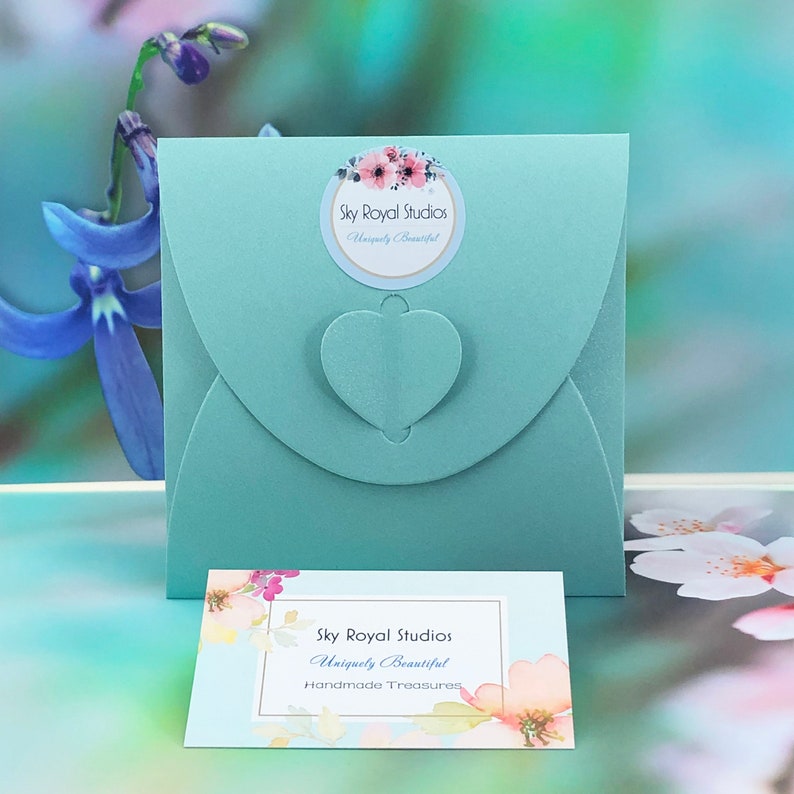 May include: A light blue envelope with a heart-shaped opening and a sticker that says "Sky Royal Studios Uniquely Beautiful". The envelope is sitting on a white surface with a business card that says "Sky Royal Studios Uniquely Beautiful Handmade Treasures".