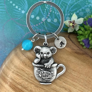 May include: Silver keyring featuring a koala bear sitting in a teacup with the text "Tea Cup Baby". The keyring also includes a turquoise bead and a silver charm with the letter "A".