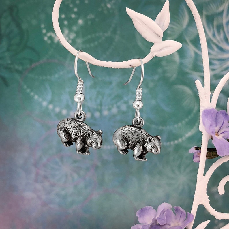 May include: A pair of silver dangle earrings featuring a detailed, three-dimensional wombat charm. The earrings are hanging from a silver hook.