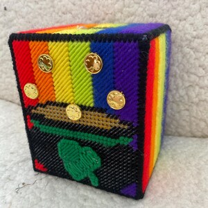 May include: A handcrafted, rainbow-colored needlepoint box with gold-colored shamrock buttons. The box features a black base with a green shamrock design. The box is a vibrant and colorful decorative item.