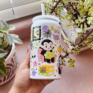 May include: White Halloween-themed tumbler with a white lid and straw. The tumbler features a cartoon vampire reading a book, surrounded by bats, spiderwebs, and autumn leaves. The text on the book reads "How to Read Politics".