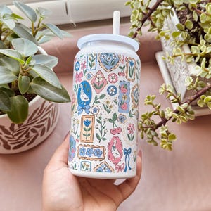 May include: White glass can-shaped tumbler with a white lid and straw. The tumbler is decorated with a repeating pattern of framed illustrations in pink, blue, and green, including flowers, birds, and bows. The tumbler is held in a hand.