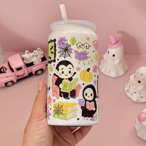 May include: A white glass tumbler with a straw featuring a colourful Halloween-themed pattern of a vampire reading a book titled How to Drink Blood Politely, a ghost reading How to Boo, a grim reaper, pumpkins, spiderwebs, and autumn leaves.