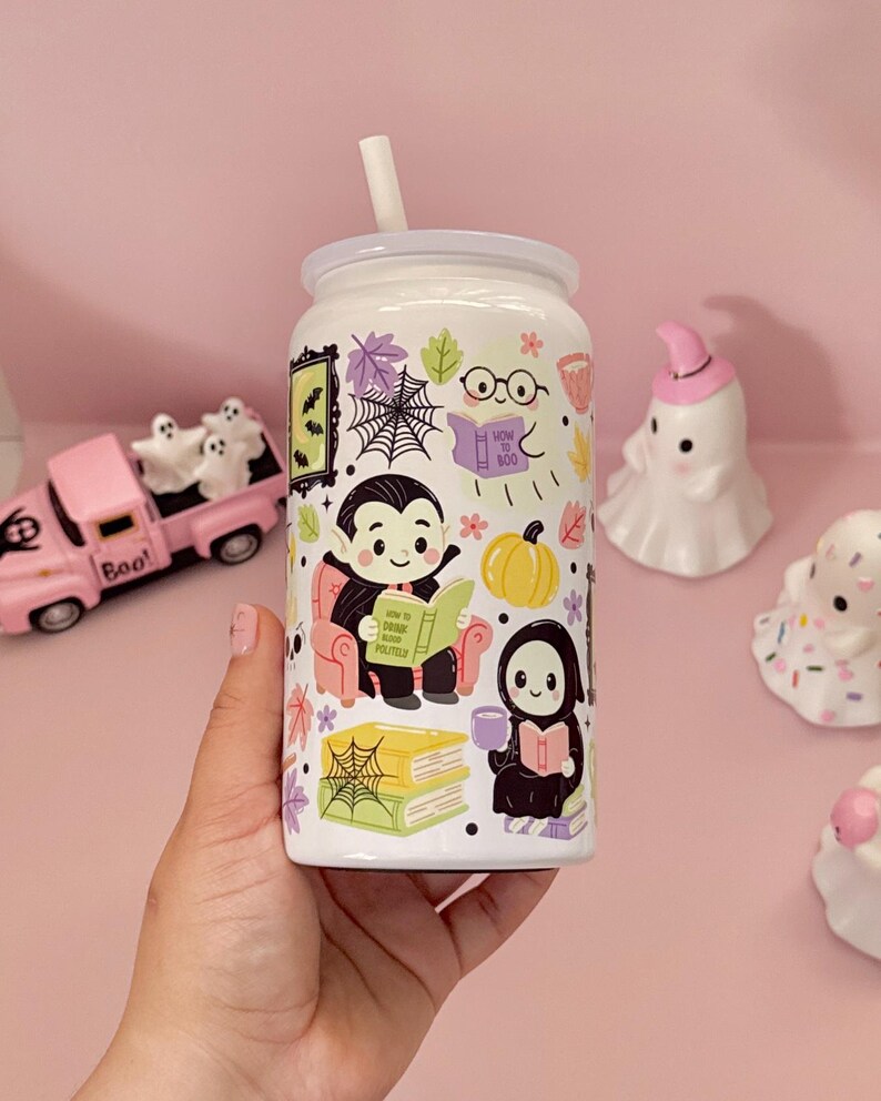 May include: A white glass tumbler with a straw featuring a colourful Halloween-themed pattern of a vampire reading a book titled How to Drink Blood Politely, a ghost reading How to Boo, a grim reaper, pumpkins, spiderwebs, and autumn leaves.
