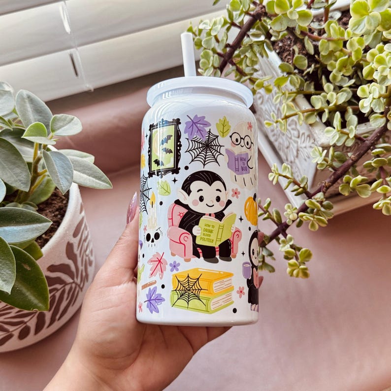 May include: White Halloween-themed tumbler with a white lid and straw. The tumbler features a cartoon vampire reading a book, surrounded by bats, spiderwebs, and autumn leaves. The text on the book reads "How to Read Politics".