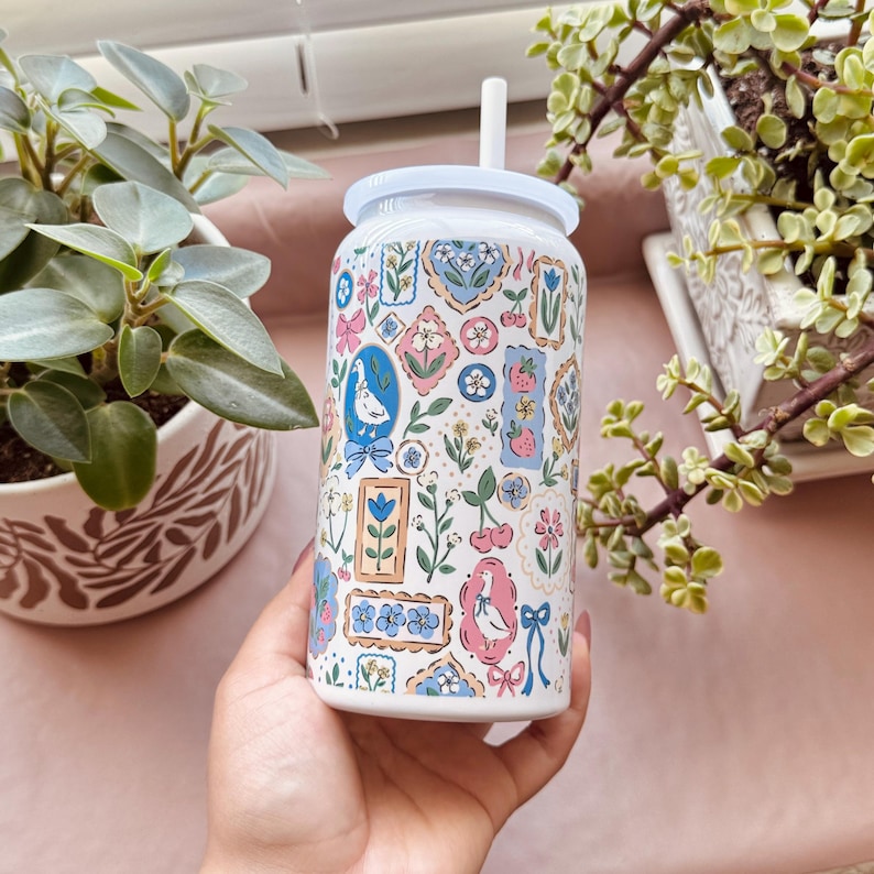 May include: White glass can-shaped tumbler with a white lid and straw. The tumbler is decorated with a repeating pattern of framed illustrations in pink, blue, and green, including flowers, birds, and bows. The tumbler is held in a hand.