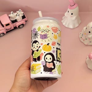 May include: A white glass can-shaped tumbler with a straw, featuring a colourful pattern of cartoon ghosts, a vampire, a grim reaper, pumpkins, books, spiderwebs, and autumn leaves. The book held by the ghost has the text How To Boo printed on it.