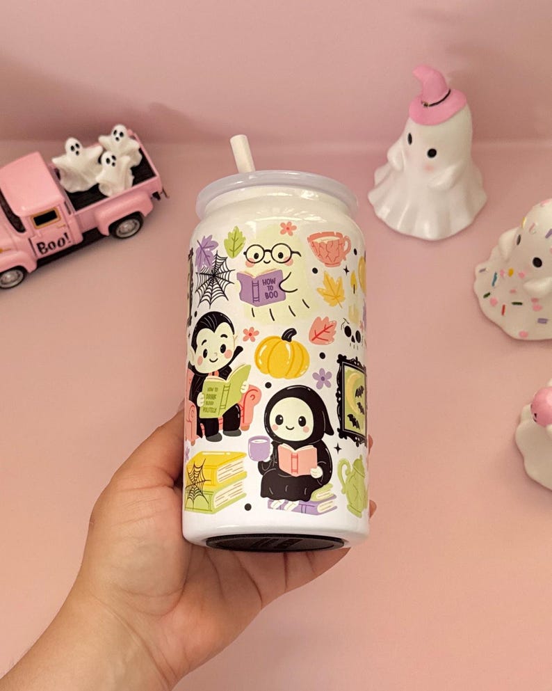 May include: A white glass can-shaped tumbler with a straw, featuring a colourful pattern of cartoon ghosts, a vampire, a grim reaper, pumpkins, books, spiderwebs, and autumn leaves. The book held by the ghost has the text How To Boo printed on it.