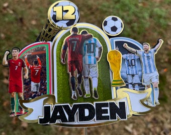 May include: A soccer-themed cake topper with a gold background and a black and white image of two soccer players, Cristiano Ronaldo and Lionel Messi, with the text "JAYDEN" in black letters. The topper also features a gold soccer ball with the number "12" on it, a gold soccer trophy, and a gold soccer net.