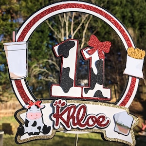 May include: A red, white, and gold glitter cake topper with a cow print number 11, a cow with a red bow, and the name Khloe. The topper also includes a red and white striped cup, a white bag with fries, and a white cup with a lid.