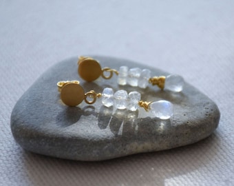 Può includere: Un paio di orecchini d'oro con un disco rotondo e un pendente di perle di pietra lunare. Gli orecchini sono su una pietra grigia.