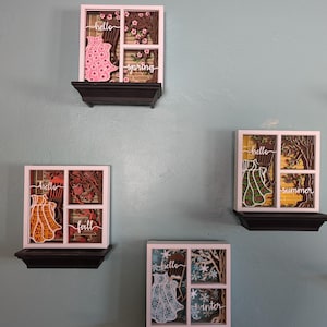 May include: Four seasonal wall art pieces, each featuring a layered design within a white frame. The art depicts a dress, trees, and seasonal text like "spring" and "winter". The pieces are mounted on black shelves against a blue wall.