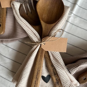 Könnte beinhalten: Ein Set aus Holzlöffel, Gabel und Pfannenwender, zusammengebunden mit einem beigefarbenen Waffelhandtuch. Löffel und Pfannenwender haben geätzten Text und ein schwarzes Herz. Ein leeres Etikett ist angebracht. Das Set befindet sich auf einer gestreiften weiß-grauen Oberfläche.