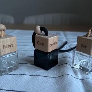 Könnte beinhalten: Drei Auto-Lufterfrischer mit Holzaufsätzen und Glasbehältern. Die Holzaufsätze haben Text und ein Herzsymbol. Einer hat "Fahrt" und ein Herz, ein anderer "Gott behüte" und der dritte "Gott beschütze". Jeder hat eine Kordel zum Aufhängen.