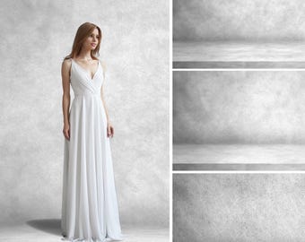 May include: A woman wearing a white sleeveless wedding dress with a V-neckline and a flowing skirt. The dress is photographed against a gray concrete background.