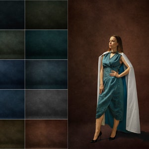 May include: A woman wearing a teal blue dress with a white cape stands in front of a brown textured background. She is looking to the left of the frame.