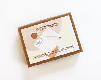 Op de afbeelding: Een bruine doos met een wit label waarop staat "50 Bedürfniskarten für Psychotherapie, Beratung und Coaching". Het label heeft een bruin en wit abstract ontwerp met een kopje koffie en een pen.