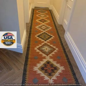 May include: A long, rectangular runner rug featuring a repeating geometric diamond pattern in shades of burnt orange, cream, dark gray, and muted blue. The rug has a dark border and is positioned centrally along a hallway floor with light walls.