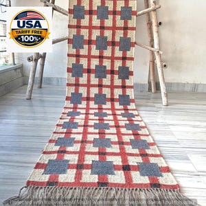 May include: A long rectangular woven runner rug featuring a grid pattern of intersecting red lines on a cream-colored background. Dark gray squares are positioned at the intersections of the red lines. The rug has fringe at the bottom edge.