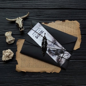 Puede incluir: Un sobre negro con un marcap&aacute;ginas blanco y gris que dice "todo comienza con un LIBRO". El marcap&aacute;ginas tiene un dise&ntilde;o de pluma negra. El sobre est&aacute; atado con una cuerda negra y est&aacute; sobre un trozo de papel viejo y amarillento.