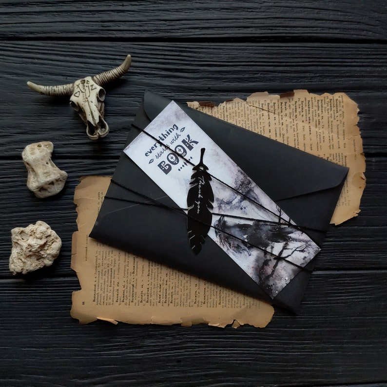 Puede incluir: Un sobre negro con un marcap&aacute;ginas blanco y gris que dice "todo comienza con un LIBRO". El marcap&aacute;ginas tiene un dise&ntilde;o de pluma negra. El sobre est&aacute; atado con una cuerda negra y est&aacute; sobre un trozo de papel viejo y amarillento.