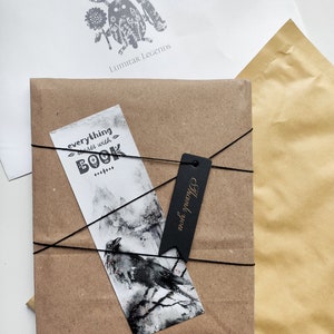 Puede incluir: Un paquete de papel marr&oacute;n atado con una cuerda y una etiqueta negra que dice "Gracias". Se puede ver un marcap&aacute;ginas con una ilustraci&oacute;n en blanco y negro de un cuervo en una rama. El marcap&aacute;ginas tiene el texto "todo comienza con un libro" en &eacute;l. El paquete est&aacute; dirigido a "Lumiar Legends".
