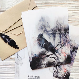 Puede incluir: Un conjunto de cuatro postales de acuarela con un cuervo negro posado en una rama frente a un paisaje monta&ntilde;oso brumoso. Las tarjetas est&aacute;n impresas con el nombre de usuario de Instagram del artista, "@KSENYA_LUMITAR", y la palabra "LUMITAR".
