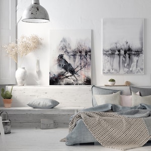 Puede incluir: Un dormitorio blanco con una cama, un banco y dos acuarelas en la pared. La pintura de la izquierda presenta un cuervo negro posado en una rama en un bosque brumoso. La pintura de la derecha es una acuarela m&aacute;s abstracta de una escena de bosque.