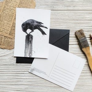 Puede incluir: Una ilustraci&oacute;n de acuarela en blanco y negro de un cuervo posado en un poste de madera. El cuervo est&aacute; mirando hacia la izquierda y tiene las alas extendidas. La ilustraci&oacute;n est&aacute; en una postal blanca con un sobre negro y pinceles en el fondo.
