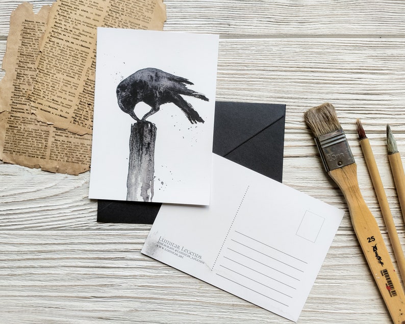Puede incluir: Una ilustraci&oacute;n de acuarela en blanco y negro de un cuervo posado en un poste de madera. El cuervo est&aacute; mirando hacia la izquierda y tiene las alas extendidas. La ilustraci&oacute;n est&aacute; en una postal blanca con un sobre negro y pinceles en el fondo.