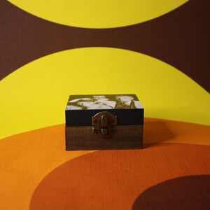 May include: A small wooden box with a black lid and a brass latch. The box has a black and white photo on the front. The box is sitting on a brown and orange background.