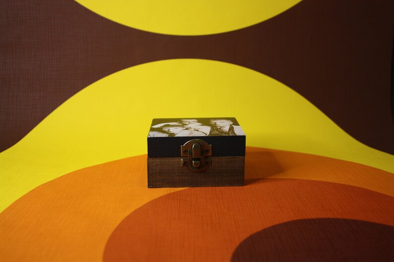 May include: A small wooden box with a black lid and a brass latch. The box has a black and white photo on the front. The box is sitting on a brown and orange background.