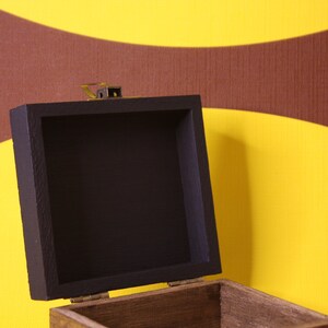 May include: An open wooden box with a black painted interior. The box has a brass latch and is sitting on a yellow and brown patterned background.